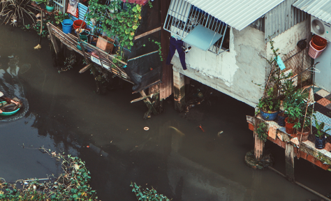 A person swims in a river that is flanked by houses with tin roofs.
